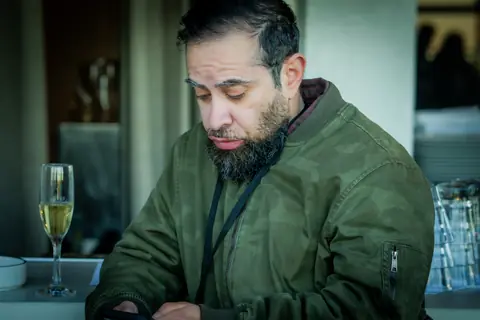 Attendee in a green jacket checking his phone with a glass of champagne