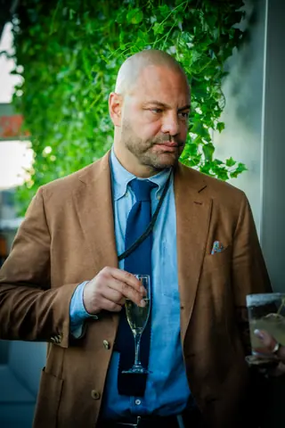 Attendee in a tan blazer with a drink beside the ivy wall