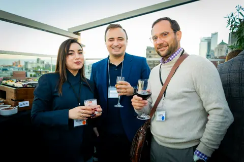 Three attendees posing with drinks on the rooftop with downtown Montreal behind them