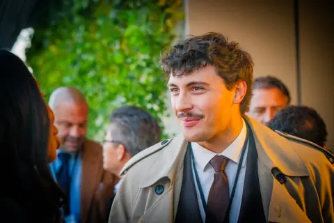 Attendee in a trench coat in conversation by the green wall