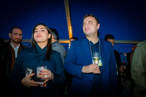 Two attendees with drinks listening to the panel discussion