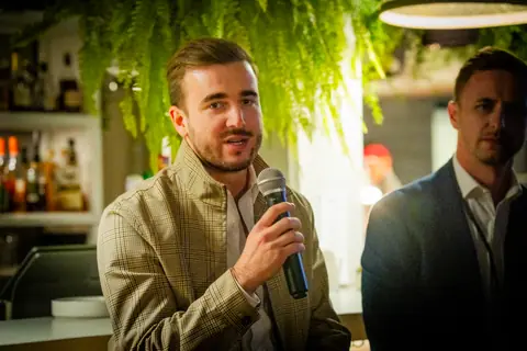 Panelist in a tan jacket speaking into a microphone