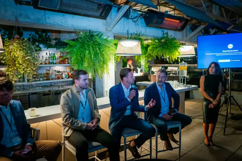 Panel of four speakers seated by the bar with a presentation screen beside them