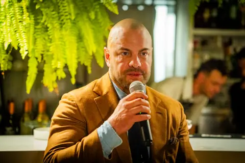 Panelist in a tan blazer speaking under the hanging ferns
