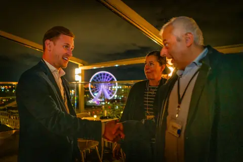 Two attendees shaking hands with the illuminated Ferris wheel behind them