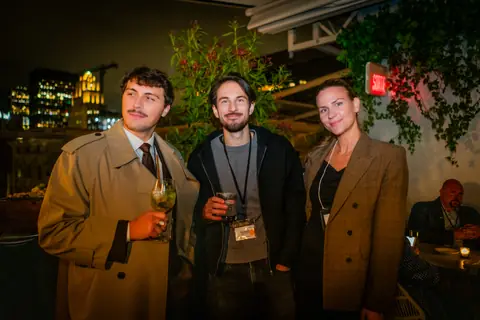 Three attendees posing together beside the ivy wall at night